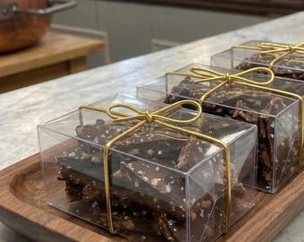 May include: Three clear rectangular plastic boxes filled with dark chocolate bark topped with sea salt. Each box is tied with a thin gold ribbon bow. The boxes are arranged in a row on a rectangular wooden tray with rounded edges and a smooth finish.