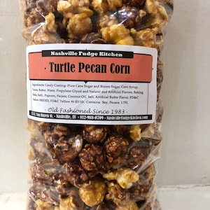 May include: A clear plastic bag filled with a mixture of caramel and chocolate popcorn. The bag has a label that reads "Nashville Fudge Kitchen" and "Turtle Pecan Corn". The label also lists the ingredients and nutritional information. The bag is sitting on a white surface.