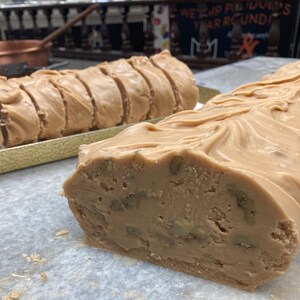 May include: A close-up of a large slab of pecan fudge, cut into slices. The fudge is a light brown color and has a smooth, glossy finish. The pecan pieces are visible throughout the fudge.