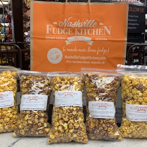 May include: Five bags of popcorn with different flavors from Nashville Fudge Kitchen. The bags are labeled with the company's name and website. The flavors are Caramel Corn, White Cheddar Popcorn, Caramel Pecan Corn, Vanilla Bean Corn, and Butter Pecan Corn.