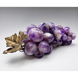 May include: A decorative cluster of polished purple amethyst stones shaped like grapes, featuring a metallic gold-toned leaf accent attached to the stem. The individual stone spheres vary in shade from light to deep violet with natural white inclusions.