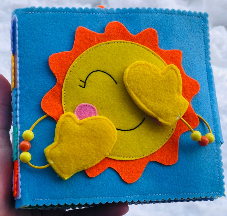 Puede incluir: Un libro de fieltro con una cara de sol sonriente con rayos naranjas. El sol sostiene dos guantes amarillos de fieltro. El libro tiene una cubierta azul con cuentas de colores.