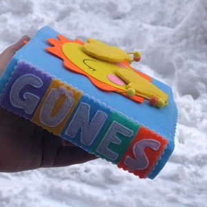 Puede incluir: Un libro de fieltro azul con la palabra "G&Uuml;NES" escrita en letras de fieltro de colores. Un sol amarillo sonriente con rayos naranjas est&aacute; en la portada.