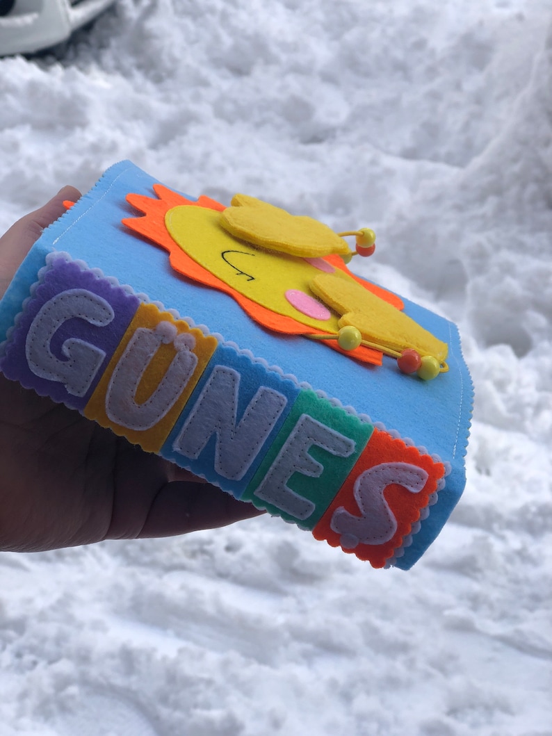 Puede incluir: Un libro de fieltro azul con la palabra "G&Uuml;NES" escrita en letras de fieltro de colores. Un sol amarillo sonriente con rayos naranjas est&aacute; en la portada.