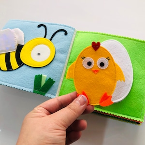 Puede incluir: Un libro de fieltro con una abeja en una p&aacute;gina y un pollito en la otra. La abeja es amarilla y negra con un c&iacute;rculo blanco en el medio. El pollito es amarillo con un coraz&oacute;n rojo en la cabeza y patas naranjas. El libro est&aacute; hecho de fieltro y tiene una textura suave.