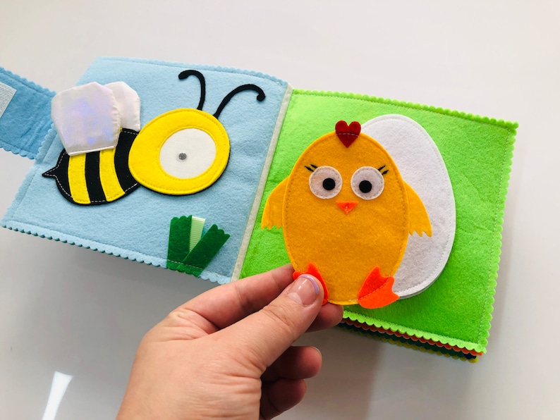 Puede incluir: Un libro de fieltro con una abeja en una p&aacute;gina y un pollito en la otra. La abeja es amarilla y negra con un c&iacute;rculo blanco en el medio. El pollito es amarillo con un coraz&oacute;n rojo en la cabeza y patas naranjas. El libro est&aacute; hecho de fieltro y tiene una textura suave.