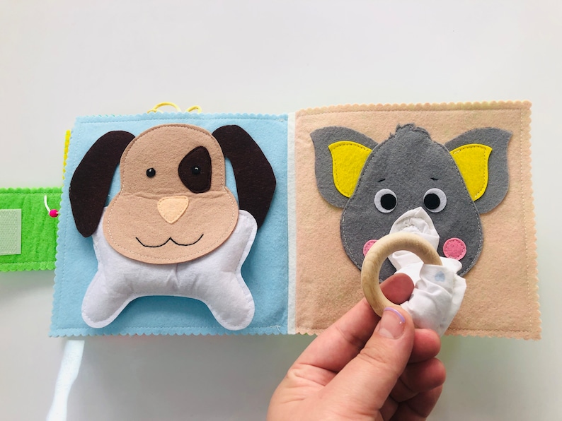 Puede incluir: Un libro de fieltro para beb&eacute;s y ni&ntilde;os peque&ntilde;os con un perro marr&oacute;n y un elefante gris en la portada. El perro tiene un hueso blanco unido a su boca y el elefante tiene un pa&ntilde;uelo blanco y un anillo de madera unido a su trompa.