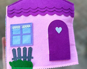 May include: A pink felt house with a purple roof, a purple door with a white heart, a blue window with a white lace trim, and a green bush with a gray fence.