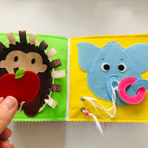 Puede incluir: Un libro de fieltro para ni&ntilde;os peque&ntilde;os con un erizo sosteniendo una manzana en una p&aacute;gina y un elefante sosteniendo una rosquilla en la otra p&aacute;gina. Las p&aacute;ginas son verdes y amarillas con costuras blancas alrededor de los bordes.