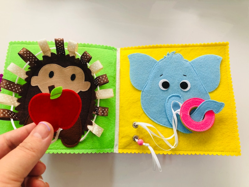 Puede incluir: Un libro de fieltro para ni&ntilde;os peque&ntilde;os con un erizo sosteniendo una manzana en una p&aacute;gina y un elefante sosteniendo una rosquilla en la otra p&aacute;gina. Las p&aacute;ginas son verdes y amarillas con costuras blancas alrededor de los bordes.