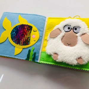 Puede incluir: Un libro de fieltro para beb&eacute;s y ni&ntilde;os peque&ntilde;os con un pez amarillo con lentejuelas arco&iacute;ris y una oveja blanca en la portada.