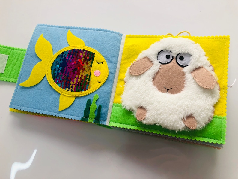 Puede incluir: Un libro de fieltro para beb&eacute;s y ni&ntilde;os peque&ntilde;os con un pez amarillo con lentejuelas arco&iacute;ris y una oveja blanca en la portada.