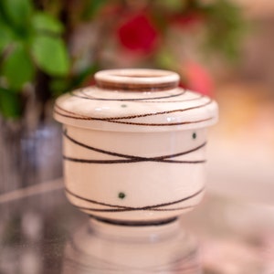 Może przedstawiać: Mały biały ceramiczny pojemnik z pokrywką. Pojemnik ma brązowy i zielony geometryczny wz&oacute;r.