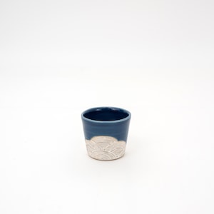 May include: A small, handmade ceramic cup with a dark blue exterior and a cream-colored, wave-like pattern near the base. The cup has a smooth, glossy finish and a simple, elegant design, perfect for beverages.
