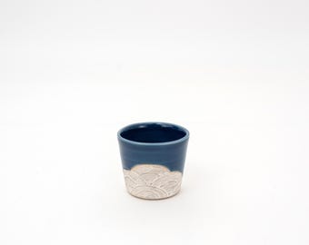 May include: A small, handmade ceramic cup with a dark blue exterior and a cream-colored, wave-like pattern near the base. The cup has a smooth, glossy finish and a simple, elegant design, perfect for beverages.