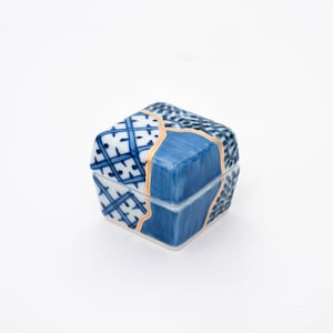 May include: A small, square ceramic box with a lid. The box is primarily blue and white with geometric patterns. Gold-colored lines highlight the repaired cracks, adding a unique aesthetic. The box is on a white background.