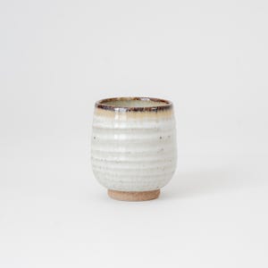 May include: A handmade ceramic cup with a textured, off-white exterior and a brown rim. The cup has a rounded shape and sits on a small, unglazed base. The design features horizontal ridges, adding to its rustic appeal. Ideal for beverages.