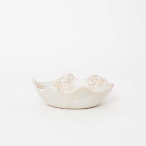 May include: A small, off-white ceramic bowl with a scalloped rim. The bowl features embossed floral designs around the edge. The bowl is a neutral color and would be suitable for serving small portions of food or as a decorative item.