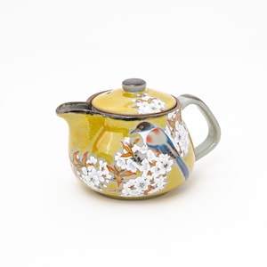 May include: A yellow ceramic teapot with a bird and floral design. The teapot has a lid and a spout. The bird is blue, red, and black, and the flowers are white.