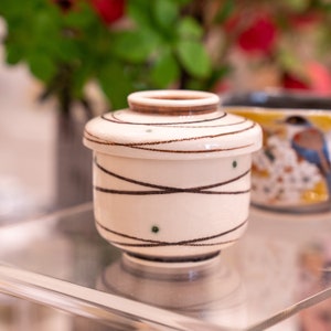 Może przedstawiać: Mały biały ceramiczny pojemnik z pokrywką. Pojemnik ma brązowy i zielony geometryczny wz&oacute;r.