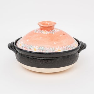 May include: A pink ceramic pot with a floral design and a black rim. The pot has two handles and a lid with a small knob on top.