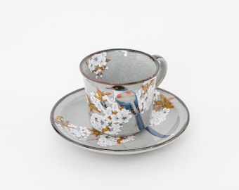 Könnte beinhalten: Ein Keramik-Tassen- und Untertassen-Set. Die graue Tasse und Untertasse zeigen ein Vogel- und Blumendesign in Weiß, Blau und Orange. Die Tasse hat einen Henkel und steht auf einer passenden Untertasse. Das Set befindet sich auf weißem Hintergrund.