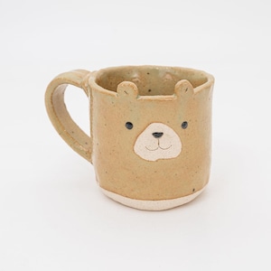 May include: A tan ceramic mug with a bear face design. The mug features a rounded body, a handle, and small bear ears at the top. The bear face has black eyes and a white snout. The mug is approximately 4 inches tall.