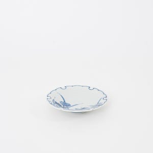 May include: A small, white ceramic dish with a scalloped edge and blue floral designs. The dish is round and has a slightly raised center. The blue design features stylized flowers and leaves.