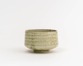 May include: A ceramic tea bowl with a light green glaze and horizontal textured lines. The bowl has a wide, rounded shape with a small footed base. Small dark speckles dot the surface, adding visual interest. The rim is a darker shade of brown.