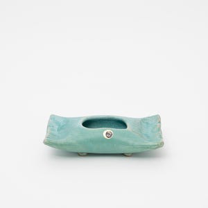 May include: A turquoise ceramic planter with a rectangular shape and a central oval opening. The planter has a textured surface and rests on three small, rounded feet. A small, round sticker is affixed to the front.