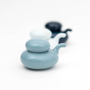 May include: A row of small, ceramic teapots in shades of blue, white, and navy. The front teapot is a light blue with a rounded lid and a curved spout. The teapots are arranged in a line, with the others slightly out of focus.