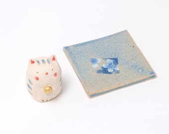 May include: A ceramic cat figurine with blue stripes and a gold accent next to a square blue coaster with a floral design. The cat has red cheeks and ears. The coaster has a textured surface.