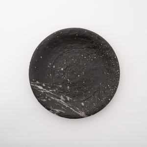 May include: A round, black ceramic plate with a textured surface. The plate has white speckles and a white paint splatter design on the lower left side. The plate is set against a white background.