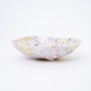 May include: A white ceramic bowl with a pink, yellow, and brown glaze. The bowl has an irregular, wavy rim and three small feet.