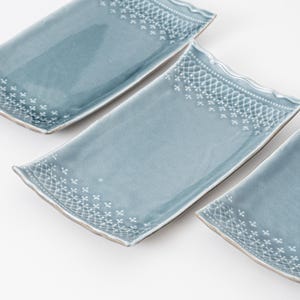 May include: Three rectangular ceramic plates with a glossy blue-grey glaze. Each plate features a delicate white lace-like pattern printed along the short edges. The edges of the plates have a subtle wavy, rippled texture and a light brown rim.