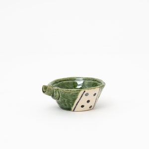 May include: Handmade ceramic bowl with a green glaze and a spout. The bowl features a cream-colored section with black dot and line details. The bowl is a unique piece of pottery.