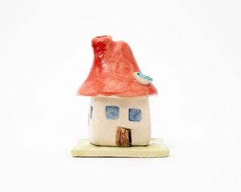 May include: A whimsical ceramic house figurine with a red roof, a chimney, and a small blue bird perched on top. The house has a cream-colored body, blue square windows, and a brown door. It sits on a green and beige base.