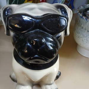 May include: A ceramic pug planter with black sunglasses, a black collar, and a cream-coloured body. The dog-shaped planter has a glossy finish and is designed to hold small plants or flowers. The pug is sitting upright, with its paws visible.