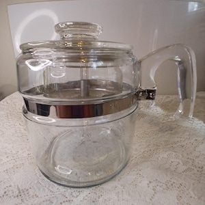 May include: A clear glass coffee pot with a glass lid and handle. The pot has a silver band around the middle and a metal filter inside. The handle is curved and attached to the side of the pot.