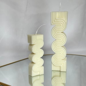 May include: Two white pillar candles with a geometric design. The candles are on a gold tray with a mirrored surface.