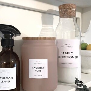 May include: Three glass bottles with cork tops and labels. The bottles are filled with white liquid. The labels read "Clean Collection Fabric Conditioner 100% Natural", "Clean Collection Laundry Pods 100% Natural", and "Clean Collection Bathroom Cleaner 100% Natural".