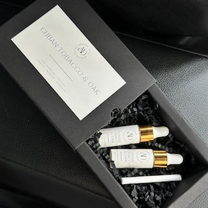 Puede incluir: Una caja negra que contiene dos pequeñas botellas blancas con tapas doradas y el texto "CUBAN TOBACCO & OAK". La caja también contiene un palito blanco y papel negro triturado.