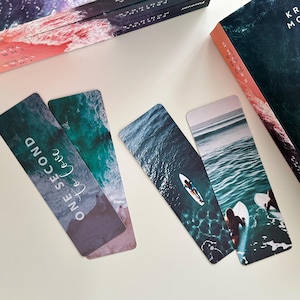 May include: Four bookmarks with ocean scenes. Two bookmarks feature a turquoise ocean with a surfer riding a wave. The other two bookmarks feature a blue ocean with surfers riding waves. One bookmark has the text "One Second to Love" printed on it.