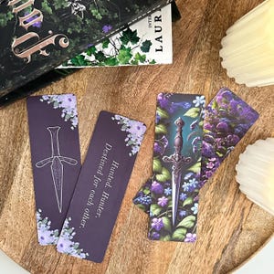 以下が含まれることがあります： 花柄と銀の短剣のイラストが描かれた、紫と緑のブックマーク3つ。ブックマークには「Hunted. Hunter. Destined for each other.」というテキストが印刷されています。