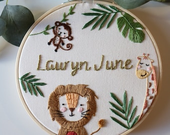 May include: A circular wooden embroidery hoop containing white fabric with hand-stitched designs. The embroidery features a brown monkey, a golden lion, and a giraffe, surrounded by green leaves. The name Lauryn June is stitched in gold thread.