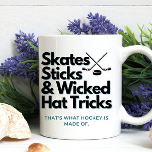 Peut inclure: Une tasse à café en céramique blanche avec du texte noir qui dit "Skates Sticks & Wicked Hat Tricks" et "That's what hockey is made of."