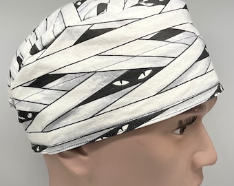 May include: A white and black patterned scrub hat with a mummy design. The hat features a white background with black bandages and black eyes.