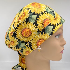 May include: A yellow and black patterned surgical scrub cap featuring a sunflower design. The cap has a tie in the back for an adjustable fit. The fabric is printed with large sunflowers, green leaves, and a black background.
