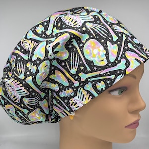 May include: A scrub cap featuring a black background with a repeating pattern of iridescent, multi-coloured human skulls, rib cages, and various bones interspersed with small white stars. The fabric has a vibrant, holographic-like finish.