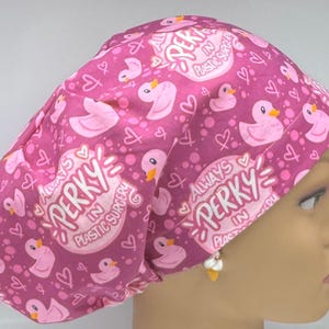 May include: A pink surgical scrub cap with a pattern of cartoon ducks, hearts, and the text "Always Perky in Plastic Surgery." The cap is made of a soft, lightweight fabric.